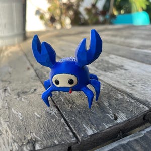 May include: A small, blue, 3D-printed crab figurine with a low-poly geometric design. The crab features two large, raised claws, multiple pointed legs, and a smooth, rounded body. It has a beige face mask with two black eyes and a small red detail.
