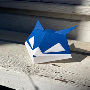 Peut inclure: Une sculpture géométrique tridimensionnelle en forme de tête de renard, composée d'une partie supérieure bleu vif et d'une partie inférieure blanche. Le design présente des lignes angulaires nettes et des oreilles pointues.