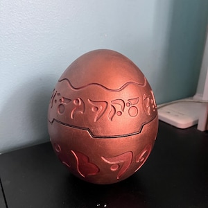 May include: A bronze-colored, egg-shaped container with intricate, raised patterns and symbols. The container is split in two halves, with a visible seam. The surface has a metallic sheen, and the design suggests a decorative or collectible item.