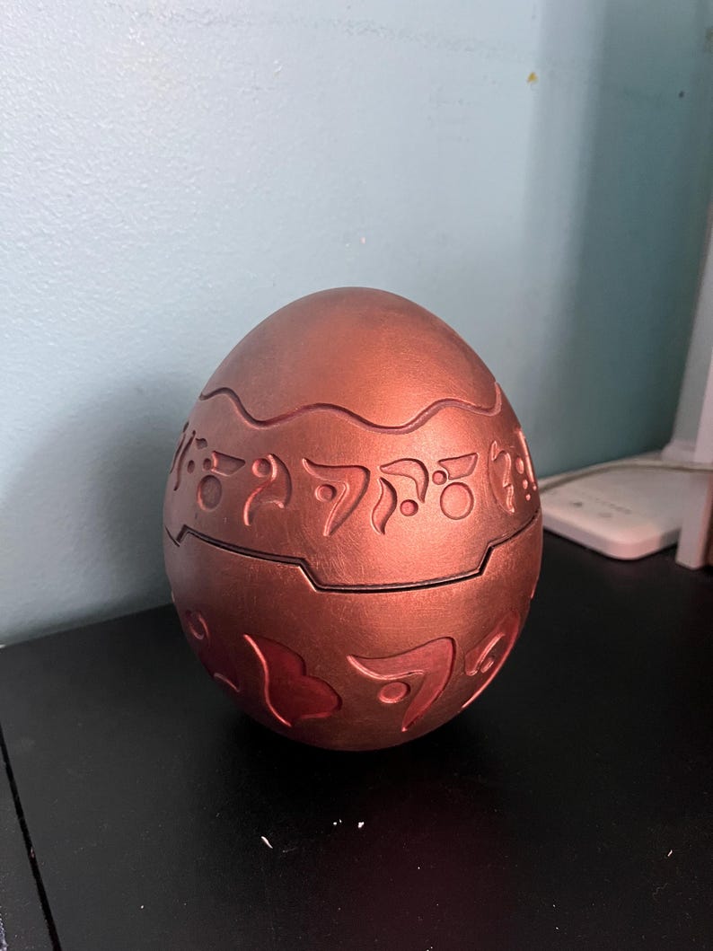 May include: A bronze-colored, egg-shaped container with intricate, raised patterns and symbols. The container is split in two halves, with a visible seam. The surface has a metallic sheen, and the design suggests a decorative or collectible item.