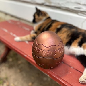 May include: A copper-colored, egg-shaped container with intricate carved designs. The container is sitting on a red bench. A calico cat is in the background.