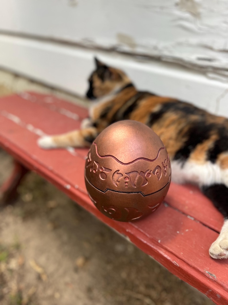 May include: A copper-colored, egg-shaped container with intricate carved designs. The container is sitting on a red bench. A calico cat is in the background.