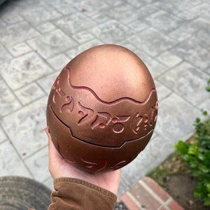 May include: A bronze-colored, egg-shaped decorative object with engraved, stylized symbols. The egg has a segmented design with a wavy line around the middle. The surface has a metallic sheen, and the object is held in a hand.