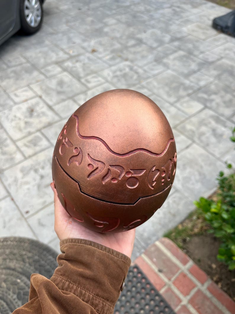 May include: A bronze-colored, egg-shaped decorative object with engraved, stylized symbols. The egg has a segmented design with a wavy line around the middle. The surface has a metallic sheen, and the object is held in a hand.