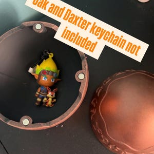 May include: A Jak and Daxter keychain figurine inside a copper-colored container. The keychain features a character with yellow hair and goggles, alongside a small orange creature. Text on a white label reads "Jak and Daxter keychain not included."
