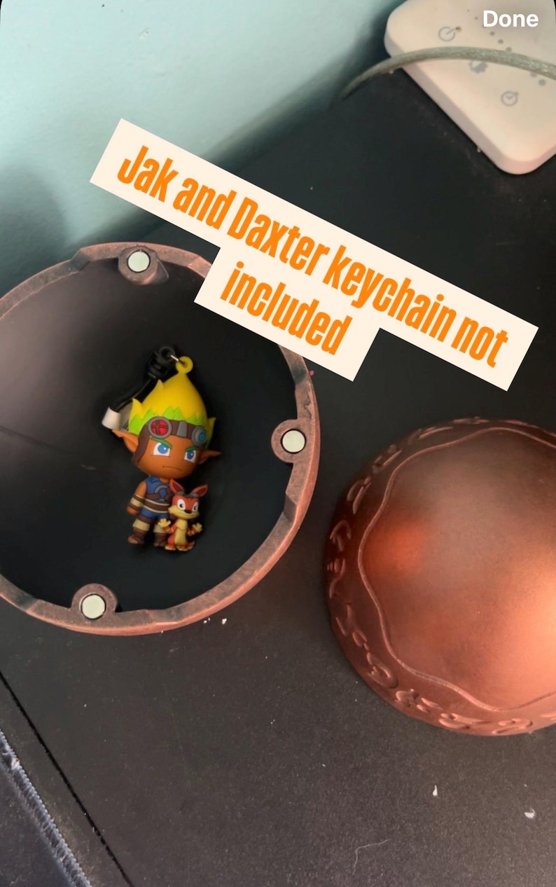 May include: A Jak and Daxter keychain figurine inside a copper-colored container. The keychain features a character with yellow hair and goggles, alongside a small orange creature. Text on a white label reads "Jak and Daxter keychain not included."