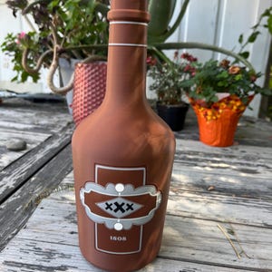 May include: A matte terracotta-colored bottle with a narrow neck and a rounded base. The front features a rectangular label with a silver-grey decorative border, three black X symbols, and the number 1808 printed in white at the bottom.