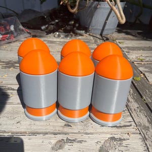 May include: A set of six cylindrical objects with a gray body and bright orange rounded caps and bases. Each object features a smooth, matte finish and a uniform design, arranged in two rows of three on a weathered wooden surface.