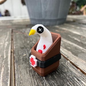 May include: A white bird figurine with a yellow beak and a black button eye, featuring red spots on its chest. The bird is positioned inside a brown, open-topped container with a black band and a white cross symbol with a red center on the front.