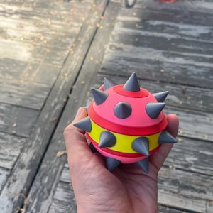 May include: A spherical toy featuring a bright pink top and bottom section with a vibrant yellow band around the center. The object is covered in multiple pointed, cone-shaped gray spikes protruding from the surface, creating a textured, spiked appearance.