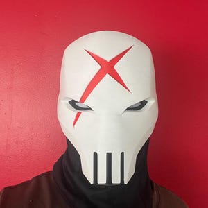 May include: A white full-face mask featuring a prominent red X symbol on the forehead, angular eye cutouts with black mesh inserts, and vertical slits at the mouth area. The mask is worn over a black neck gaiter and a plain brown shirt.