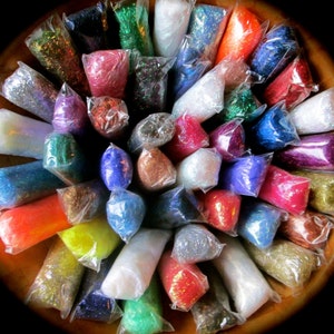 May include: A collection of small, clear plastic bags filled with colorful glitter. The glitter comes in a variety of colors, including red, blue, green, and gold. The bags are arranged in a circular pattern, creating a vibrant display.