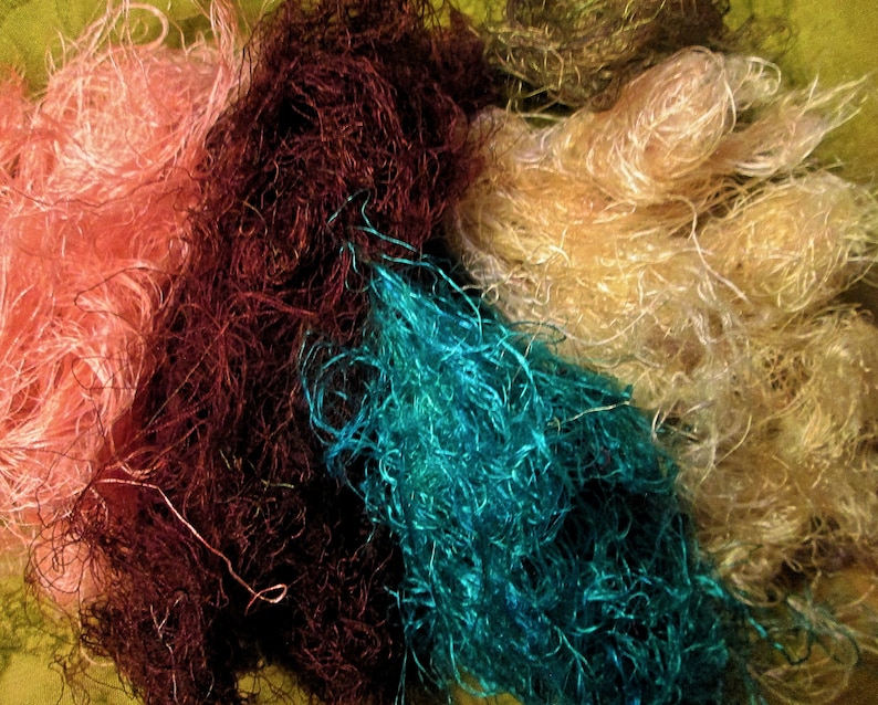 May include: A collection of colorful, fluffy yarn fibers in shades of pink, brown, teal, and cream. The fibers are loose and tangled, creating a textured and visually appealing display.