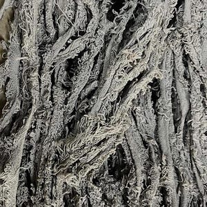 May include: Close-up of a gray, textured fabric with a frayed, fringed appearance. The fabric is composed of numerous strands, creating a dense, layered effect. The material appears soft and possibly handmade.