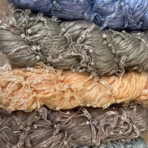 May include: Close-up of several skeins of yarn in various colors, including blue, gray, peach, and brown. Each skein is adorned with delicate lace trim and small, white faux pearls, creating a textured and decorative effect. Ideal for crafting projects.