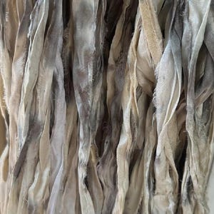 May include: Close-up of a collection of fabric strips in varying shades of cream, gray, and brown. The strips are textured and appear to be made of a soft material, possibly silk or a similar fabric. The overall effect is of a flowing, layered textile.