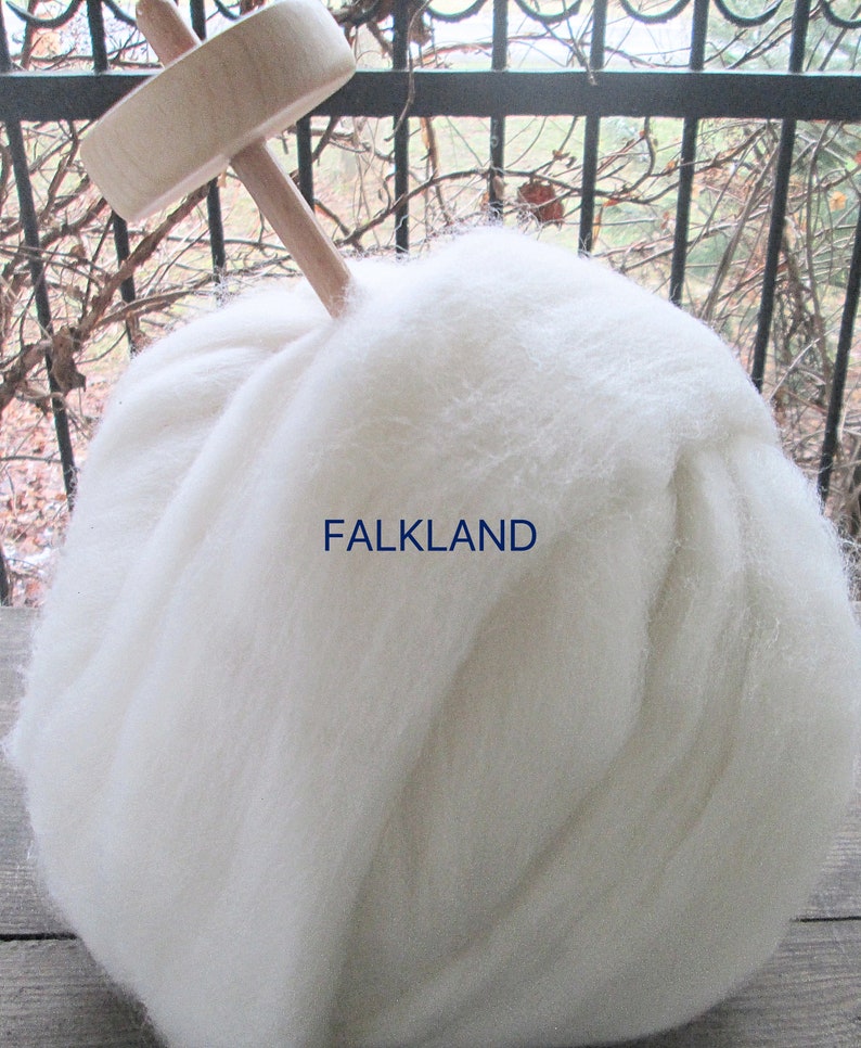 May include: A large ball of white wool with a wooden spindle inserted in the top. The text "FALKLAND" is printed in blue on the wool. The wool appears soft and fluffy, suitable for spinning or crafting.