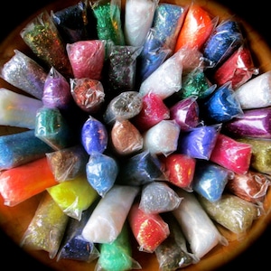 May include: Assortment of colorful glitter in individual clear plastic bags. The glitter is arranged in a circle on a wooden surface.