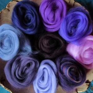 May include: A collection of colorful wool roving in shades of blue, purple, and pink, arranged in a circular pattern within a decorative, gold-rimmed bowl. The wool appears soft and fluffy, ideal for fiber art projects.