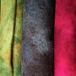 May include: Close-up of three rolls of colorful felt fabric. The rolls display shades of green, dark blue-gray, and vibrant magenta. The felt has a soft, textured appearance, suggesting it is made of wool or a similar material. The image highlights the rich colors and textures.