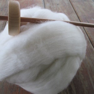 May include: A wooden drop spindle with a gold hook rests on a fluffy cloud of white wool roving. The spindle has a round whorl and a long, slender shaft, perfect for hand spinning yarn. The wool is soft and inviting.