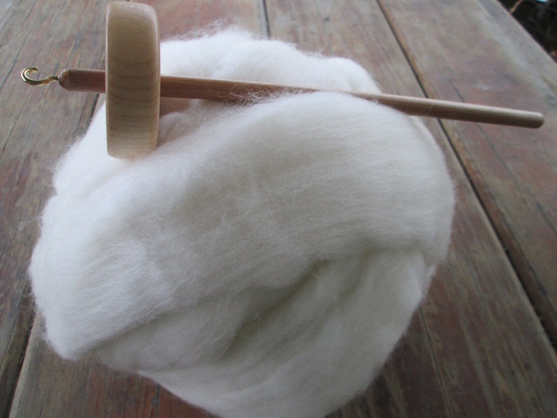 May include: A wooden drop spindle with a gold hook rests on a fluffy cloud of white wool roving. The spindle has a round whorl and a long, slender shaft, perfect for hand spinning yarn. The wool is soft and inviting.