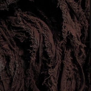 May include: A dark brown fabric with a heavily distressed and frayed texture. The material consists of numerous thin, interwoven strips that create a dense, layered appearance with loose threads and ragged edges throughout the surface.