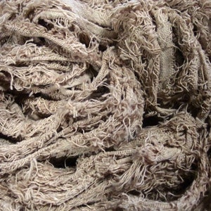 May include: Close-up of a tangled mass of beige fabric with a rough, textured appearance. The fabric appears to be made of woven strips with frayed edges, creating a layered, organic look. The material is tightly wound.