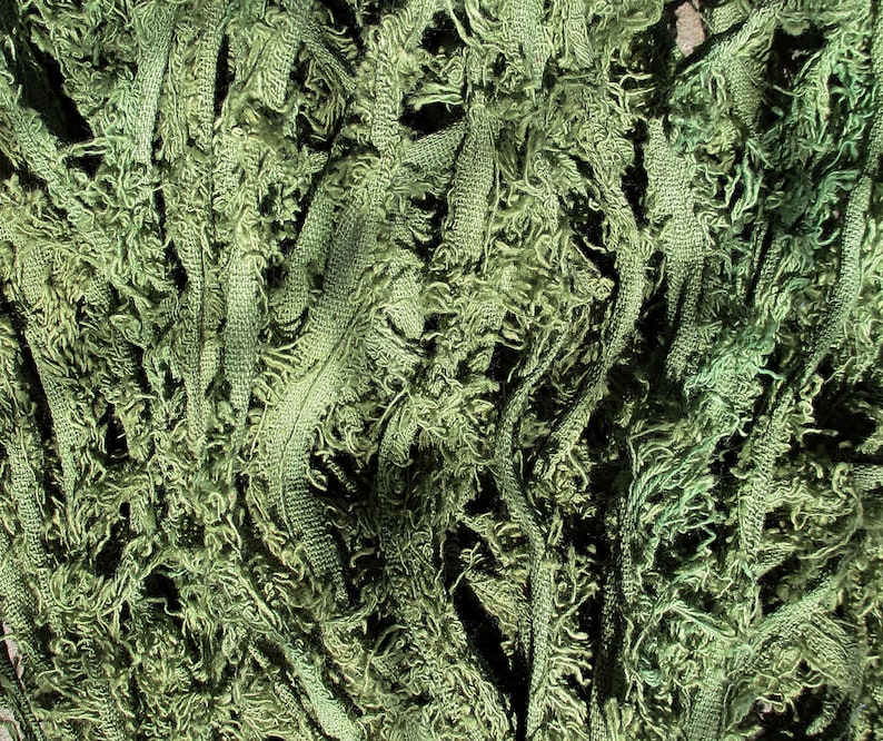 Puede incluir: Primer plano de una tela verde oliva texturizada. El material parece estar hecho de hilos entrelazados con una textura deshilachada y con flecos. La tela tiene una apariencia densa y en capas, con diferentes tonos de verde.