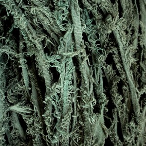 Puede incluir: Primer plano de una tela texturizada de color verde salvia. La tela parece estar hecha de tiras con bordes deshilachados, creando una apariencia en capas y algo desgastada. La textura es densa y visualmente interesante.