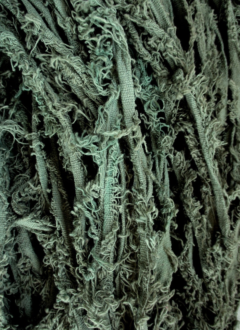 Puede incluir: Primer plano de una tela texturizada de color verde salvia. La tela parece estar hecha de tiras con bordes deshilachados, creando una apariencia en capas y algo desgastada. La textura es densa y visualmente interesante.