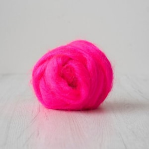 May include: A vibrant, neon pink wool roving, tightly coiled into a round shape. The fiber is soft and fluffy, perfect for needle felting or other fiber art projects. The word "Shocking" is in the bottom right corner.