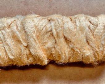May include: A close-up of a bundle of light yellow fiber, possibly silk or a similar material. The fibers are tightly packed and appear to be twisted or braided together, creating a textured surface. The color is a soft, warm tone.