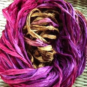 May include: A bundle of hand-dyed silk ribbon in shades of pink, purple, and brown. The ribbon is arranged in a loose coil, with the ends of the ribbon trailing out.