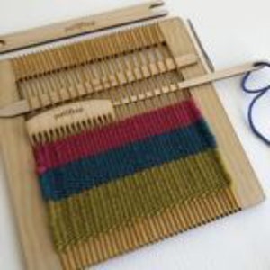 May include: A wooden frame loom with a woven textile featuring horizontal stripes in magenta, teal, and olive green. The set includes a wooden weaving comb and shuttle, both branded with the word Purl & Loop, alongside a length of blue yarn.