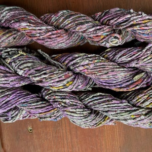 May include: Three skeins of purple and white recycled paper yarn. The yarn is made from shredded paper and is twisted together to create a rope-like texture.