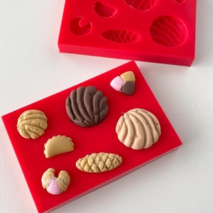 May include: Two red silicone molds for making pastries. The top mold has six cavities, including a round shape with a swirl design, a smaller round shape, a crescent shape, a leaf shape, and two other shapes. The bottom mold has eight cavities, including a round shape with a swirl design, a smaller round shape, a crescent shape, a leaf shape, and three other shapes.