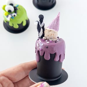 May include: Three miniature cakes decorated for Halloween. One is green with googly eyes, one is black with purple and white sprinkles, and one is purple with a pink cone and black and white striped candy.