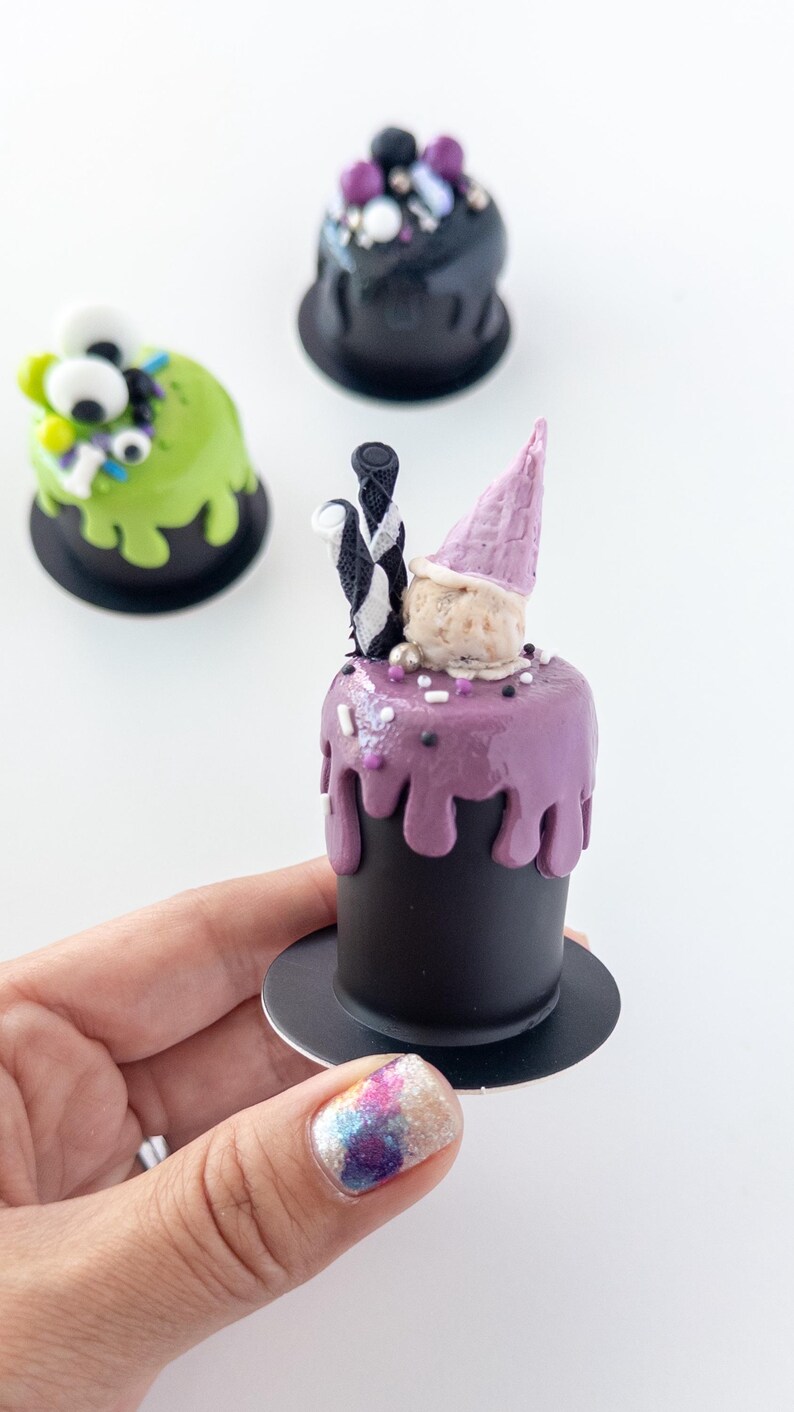 May include: Three miniature cakes decorated for Halloween. One is green with googly eyes, one is black with purple and white sprinkles, and one is purple with a pink cone and black and white striped candy.