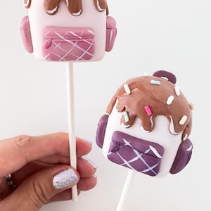 May include: Two cake pops decorated as backpacks with brown icing and colorful sprinkles. One backpack is pink and the other is purple.
