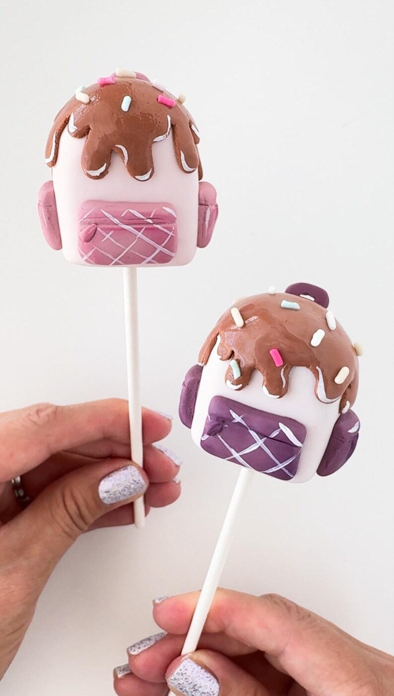 May include: Two cake pops decorated as backpacks with brown icing and colorful sprinkles. One backpack is pink and the other is purple.