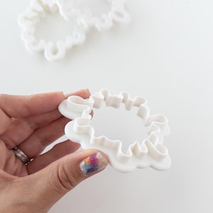 May include: Two white plastic cookie cutters shaped like a flower with scalloped edges. The larger cutter is in the background and the smaller cutter is being held in a hand.