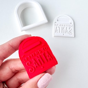 May include: A red cookie cutter in the shape of a mailbox with the words "LETTERS TO SANTA" embossed on it. A white arch-shaped cutter and a white stamp with the same mailbox design are also visible. The items are likely for holiday baking.