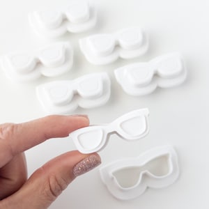 May include: White plastic eyeglasses-shaped cookie cutters. One cutter is held by a hand with a glittery nail polish. Several other cutters are scattered on a white surface. The cutters are for making cookies or other treats.