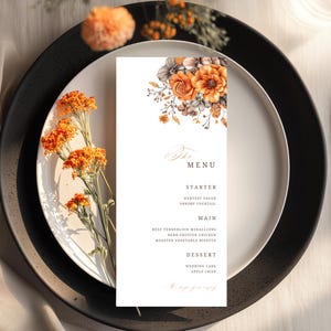 May include: A formal table setting with a white plate, black charger, and black silverware. A menu card with floral accents and menu details is placed on the plate. Orange flowers and a glass of water complete the elegant arrangement.