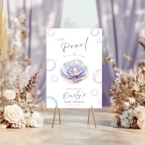 Peut inclure: Un panneau blanc sur un chevalet doré avec le texte "a little Pearl is on the way" et "Welcome to Emily's Baby Shower". Le panneau présente une fleur et des bulles à l'aquarelle.