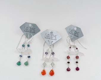 May include: Three pairs of dangle earrings on diamond-shaped cards labeled Salome Jewelry Designs. Each pair features silver-tone hooks, clear quartz points, and small faceted gemstone beads in green, orange, and deep pink color palettes.