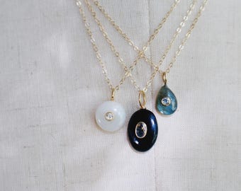 May include: Three gold chain necklaces featuring distinct pendants: a circular white stone, an oval black stone, and a teardrop-shaped blue-green stone. Each pendant is centered with a small, clear, round gemstone set in a gold bezel.