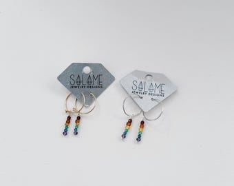 May include: Two pairs of hoop earrings on gray diamond-shaped display cards labeled Salame Jewelry Designs. One pair is gold-toned and the other is silver-toned. Each hoop features a dangling vertical strand of small, multi-colored beads.