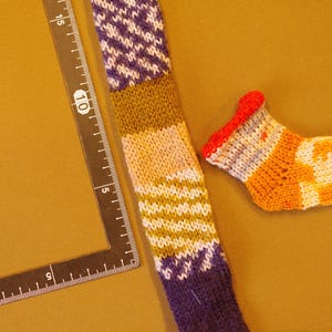 K&ouml;nnte beinhalten: Eine bunte gestrickte Socke mit einem lila, gr&uuml;nen und wei&szlig;en Muster. Eine kleinere, gestreifte Socke mit orange, wei&szlig;en und roten Streifen ist ebenfalls abgebildet.